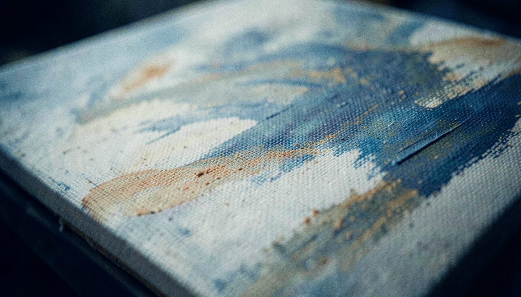 A softly lit close-up of a textured canvas with broad brushstrokes in muted blues, ochres, and creams, evoking a quiet studio atmosphere